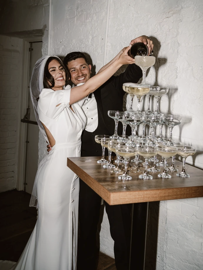 Bridal couple pouring Champagne on champagne tower at wedding reception Photo by Jessica Lily